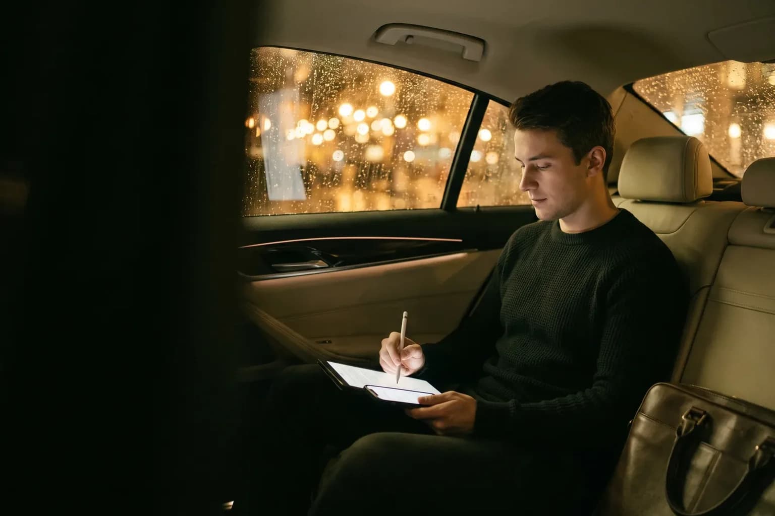 A young man signs a document on a tablet in the back seat of a car at night, city lights blurred in the rain-streaked window