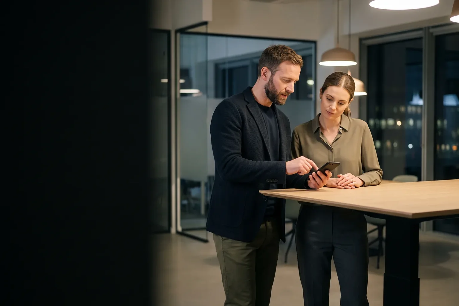 Two colleagues stand at a high table in an open-plan office at night; he signs on a smartphone while she looks on